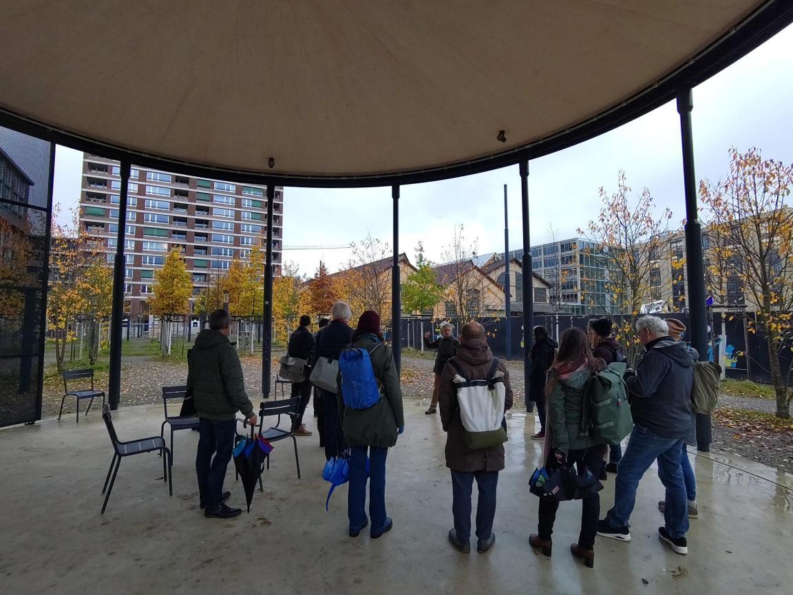 Personen stehen in einem Pavillon mit Stühlen, rundherum sind grössere und kleinere Gebäude