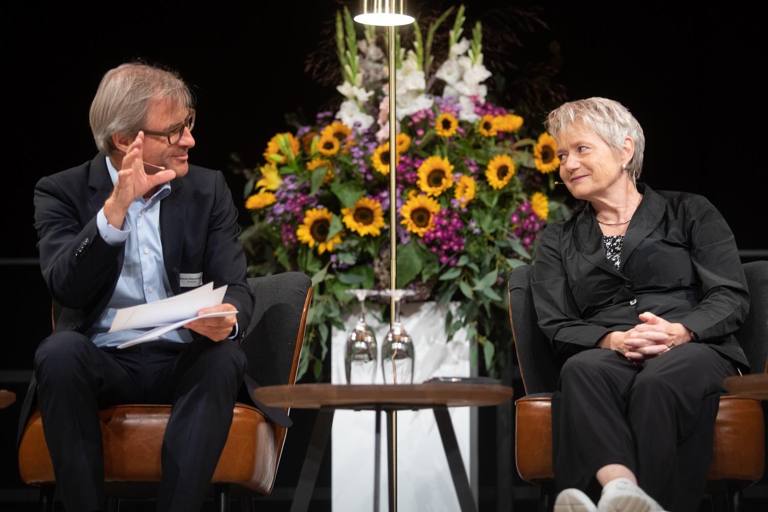 Bühnenszene. Moderator Michael Emmenegger diskutiert mit Regierungspräsidentin Jacqueline Fehr. Im Hintergrund ist ein grosses Blumengesteck zu sehen.