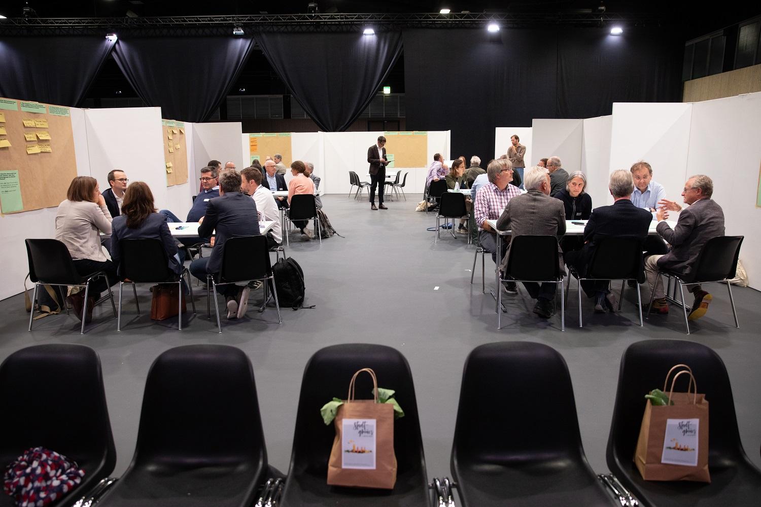 Gruppenarbeit am Klimadialog 2021: In Kleingruppen sitzen die Teilnehmenden um Tische. Am Kopf der Tische sind Trennwände mit Flipcharts aufgestellt. Auf diesen notieren die Workshop-Gruppen ihre Erkenntnisse auf bunten Post-it-Zetteln.