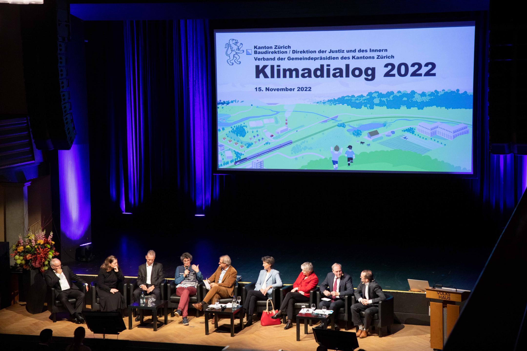 Die neun Personen aus Politik, Verwaltung und Fachthemen sitzen nebeneinander in Sesseln auf einer Bühne. Im Hintergrund ist die Titelfolie der Präsentation mit der Aufschrift Klimadialog 2022 zu sehen