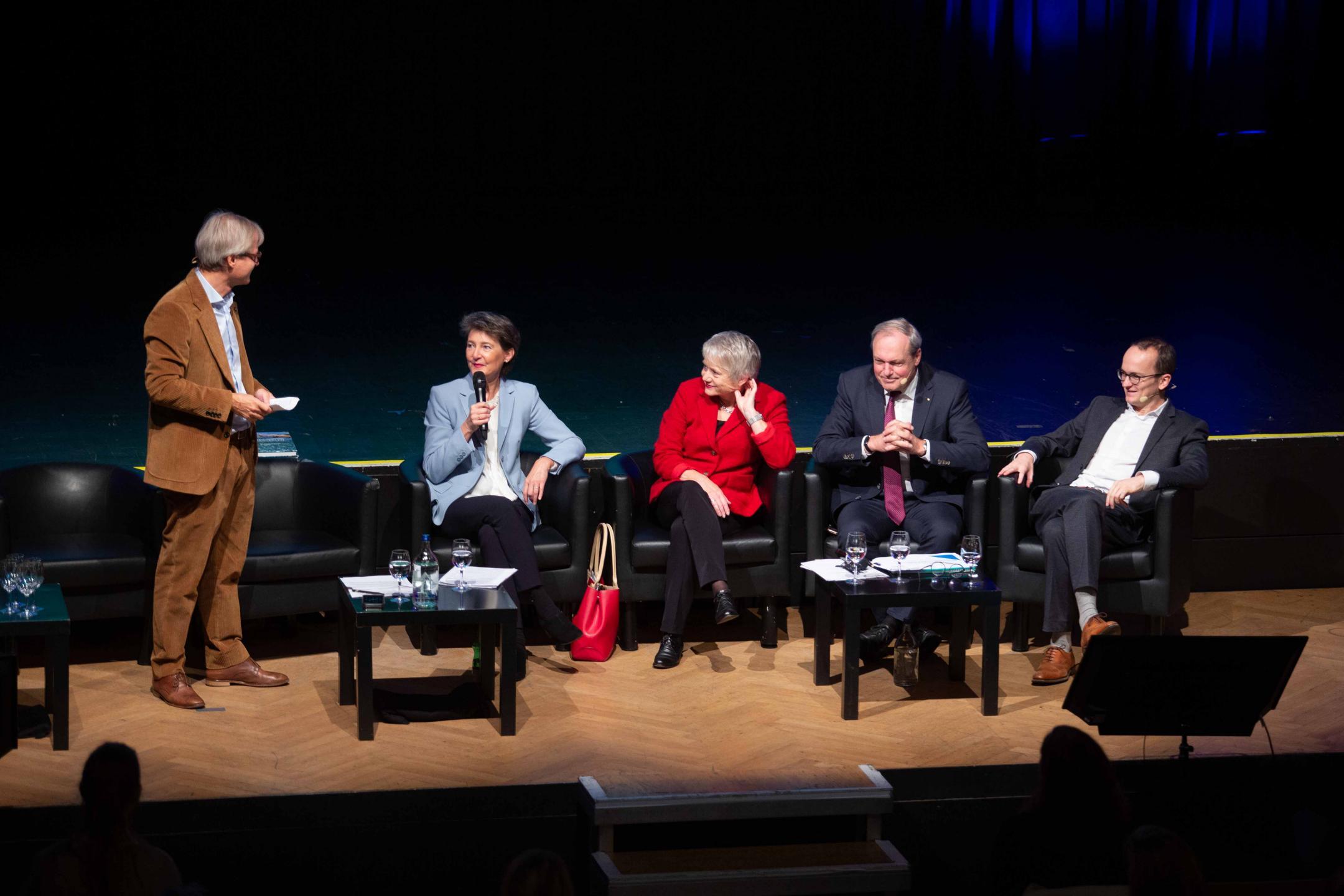 die genannten sitzen in einem Sessel auf der Bühne, der Moderator steht