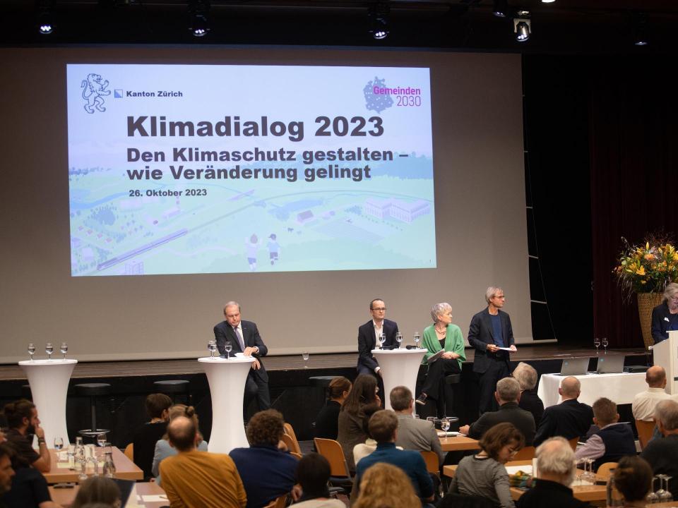 Stadtpräsidentin steht am Rednerpult, Moderator sowie Gastgebende hören von Bühne aus zu