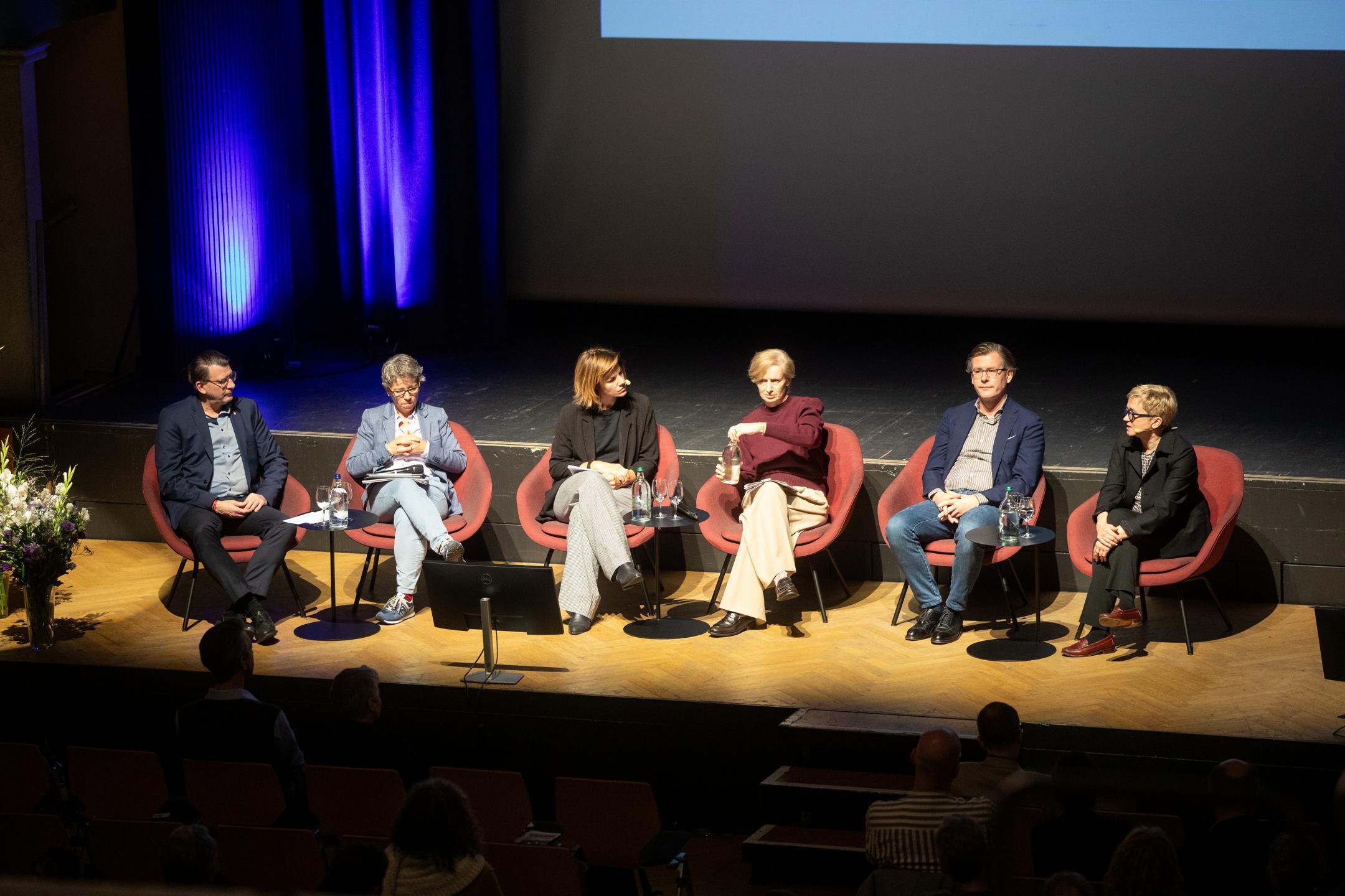 Die Podiumsgäste sitzen in einer Reihe auf der Bühne.