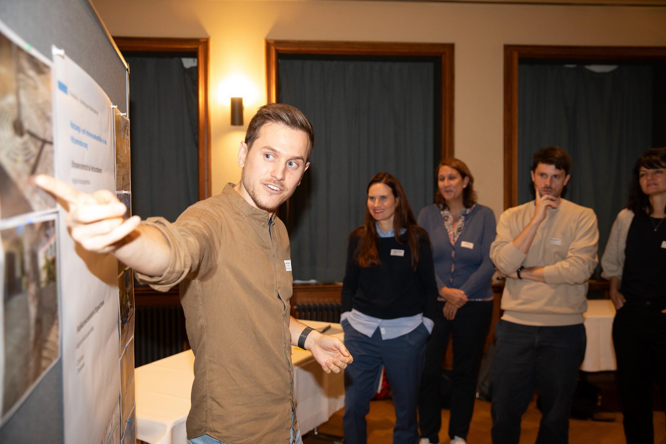 Eine Person zeigt etwas auf einem Plakat, mehrere Personen hören interessiert zu.