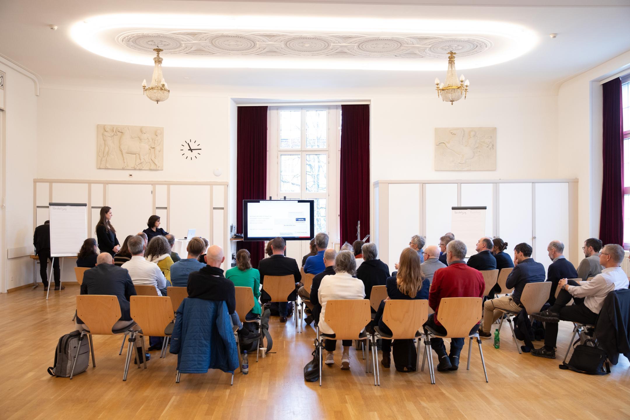 Mehrere Personen von hinten, sitzen auf Stühlen und hören Vortrag zu.