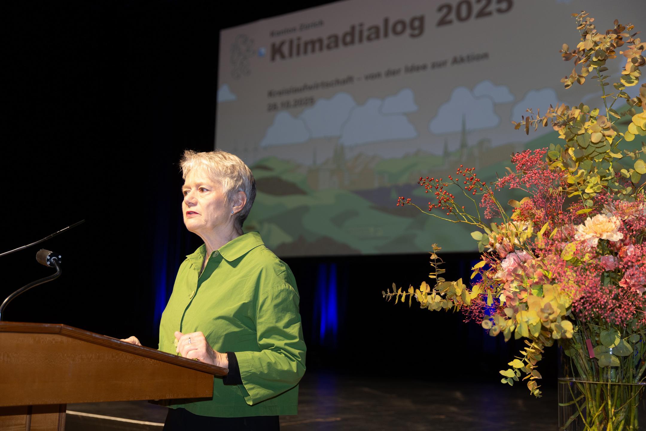 RR Jacqueline Fehr an Rednerpult, rechts von ihr ein Blumenbouqet, im Hintergrund Präsentationsfolie mit Aufschrift Klimadialog