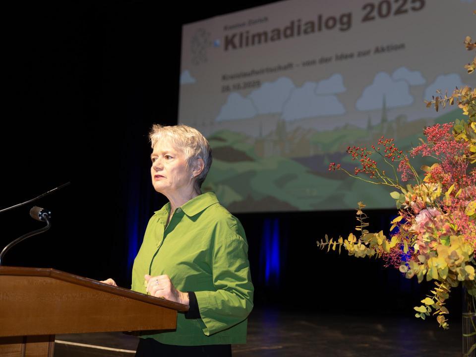 RR Jacqueline Fehr an Rednerpult, rechts von ihr ein Blumenbouqet, im Hintergrund Präsentationsfolie mit Aufschrift Klimadialog