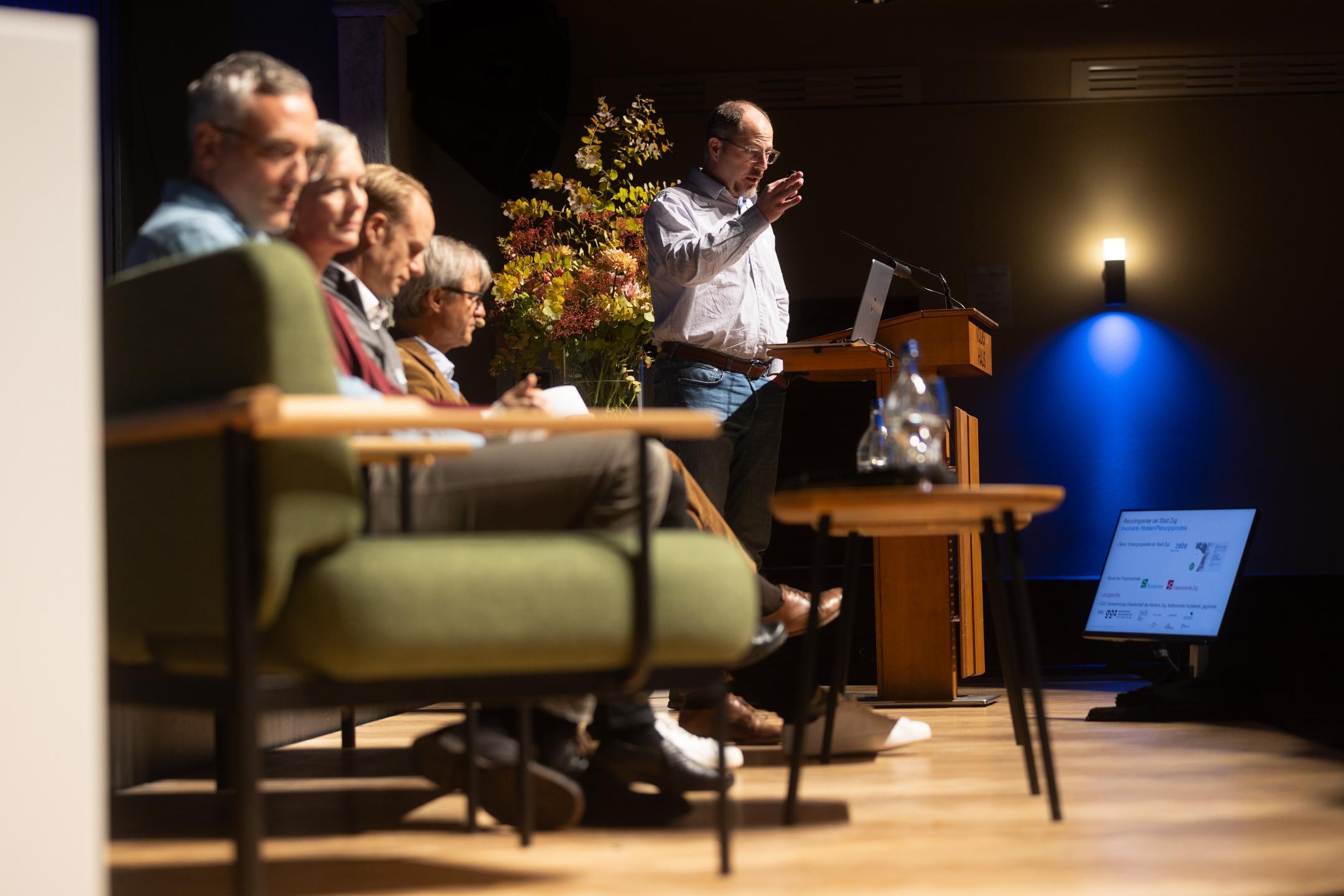 Fokus liegt auf Person, die Referat an Rednerpult hält, links im Bild sitzen weitere Personen auf der Bühne und hören zu.