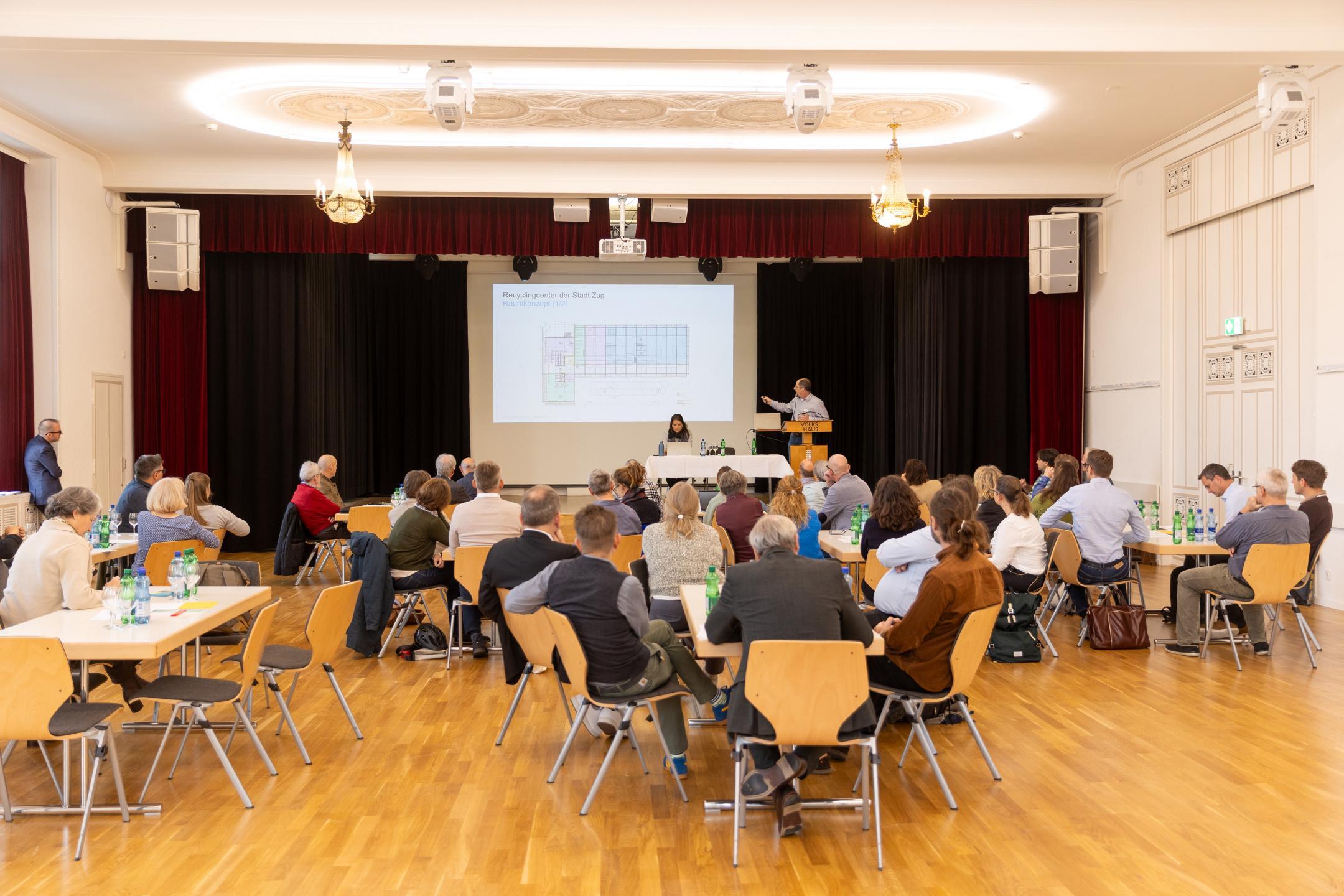 Saal mit Personen, die an Tischen verteilt sitzen und einer Präsentation zuhören
