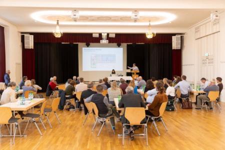 Saal mit Personen, die an Tischen verteilt sitzen und einer Präsentation zuhören