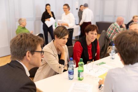 Personen sitzen an einem Tisch, weitere Personen im Hintergrund