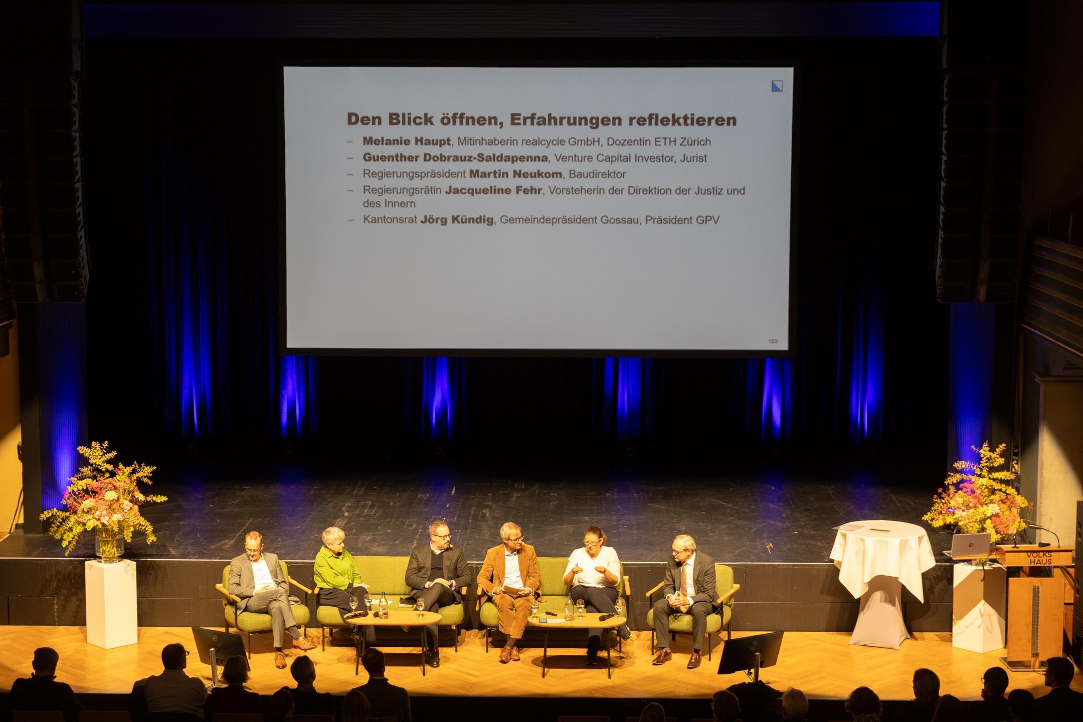 Sechs Personen sitzen auf Podium, eine eingeblendete Folie benennt die Podiumsgäste