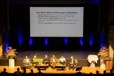 Sechs Personen sitzen auf Podium, eine eingeblendete Folie benennt die Podiumsgäste