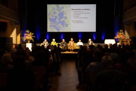 Publikum, Podiumsgäste auf der Bühne, Folie im Hintergrund zeigt, Kantonsumriss, blau eingefärbt sind die Gemeinden, die sich in Arbeitsgruppen engagieren