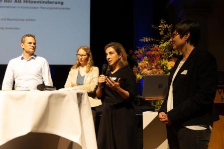 Personen stehen um Stehtisch, eine Person spricht ins Mikrofon