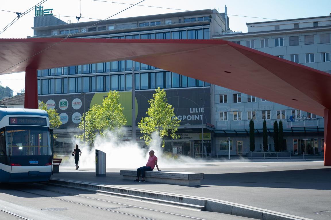 Der Stadtplatz mit seinem typischen roten Dach, ein Tram hält an, ein Mann sitzt auf einer Bank aus Stein, eine Joggerin läuft am Wassernebel vorbei
