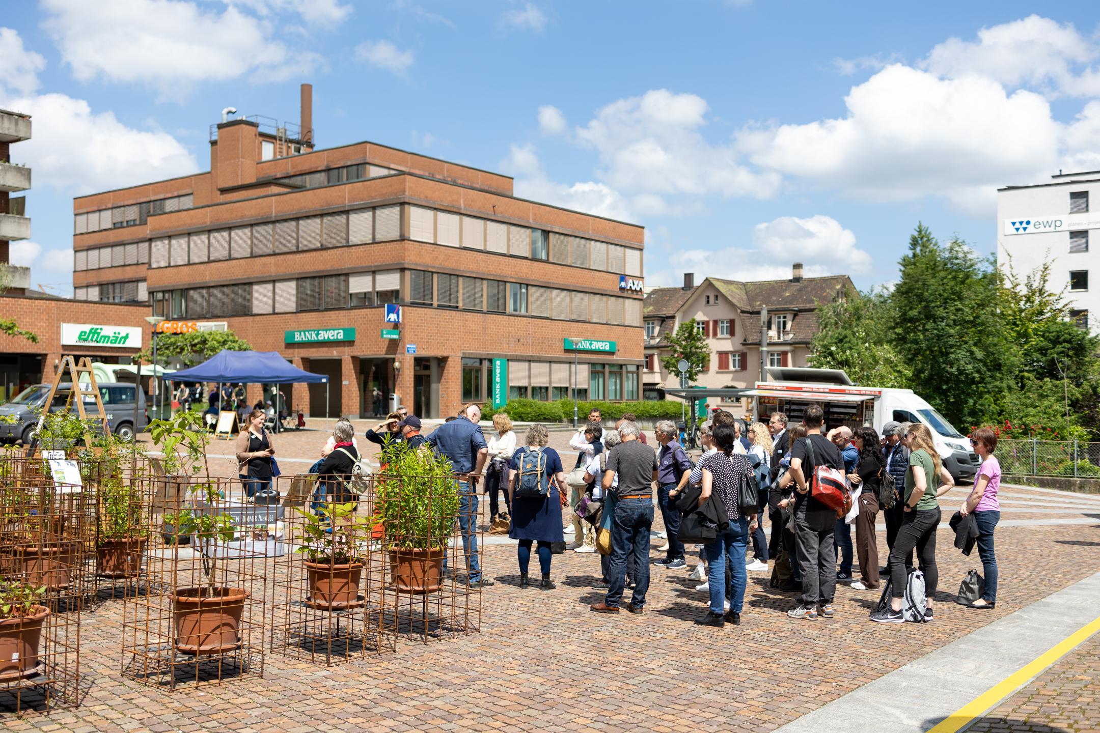 Teilnehmende stehen auf dem gepflasterten Märtplatz zusammen, links eine temporäre Neophyten-Ausstellung, im Hintergrund Backsteingebäude