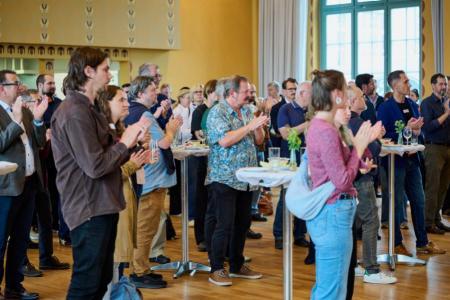 Personen in einem Raum an Stehtischen, sie applaudieren