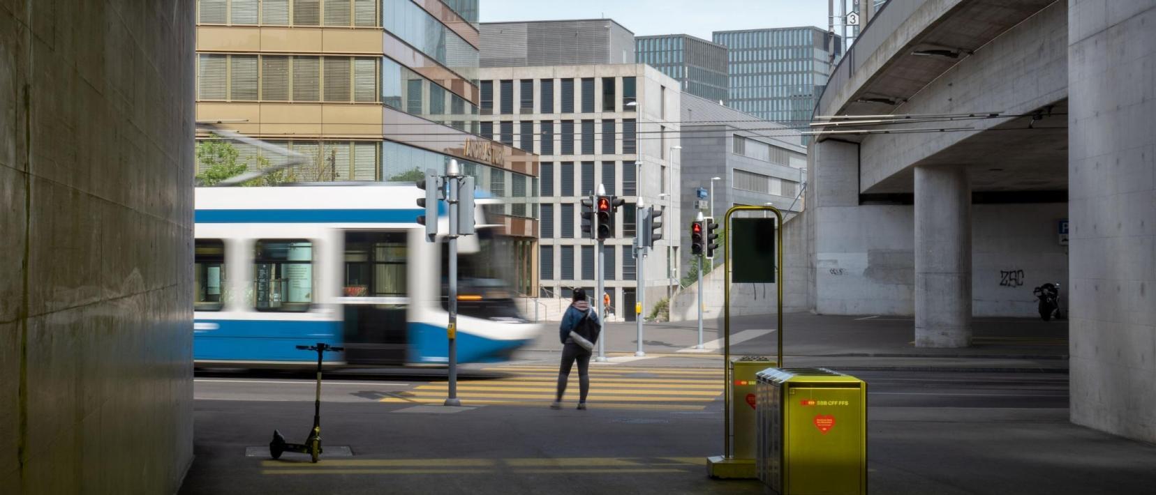 Städtische Szene mit einem Tram, das von links nach rechts fährt, während eine Person am Strassenrand wartet. Im Hintergrund sind moderne Gebäude zu sehen. Das Bild ist aus der Perspektive unter einer Betonüberdachung aufgenommen.