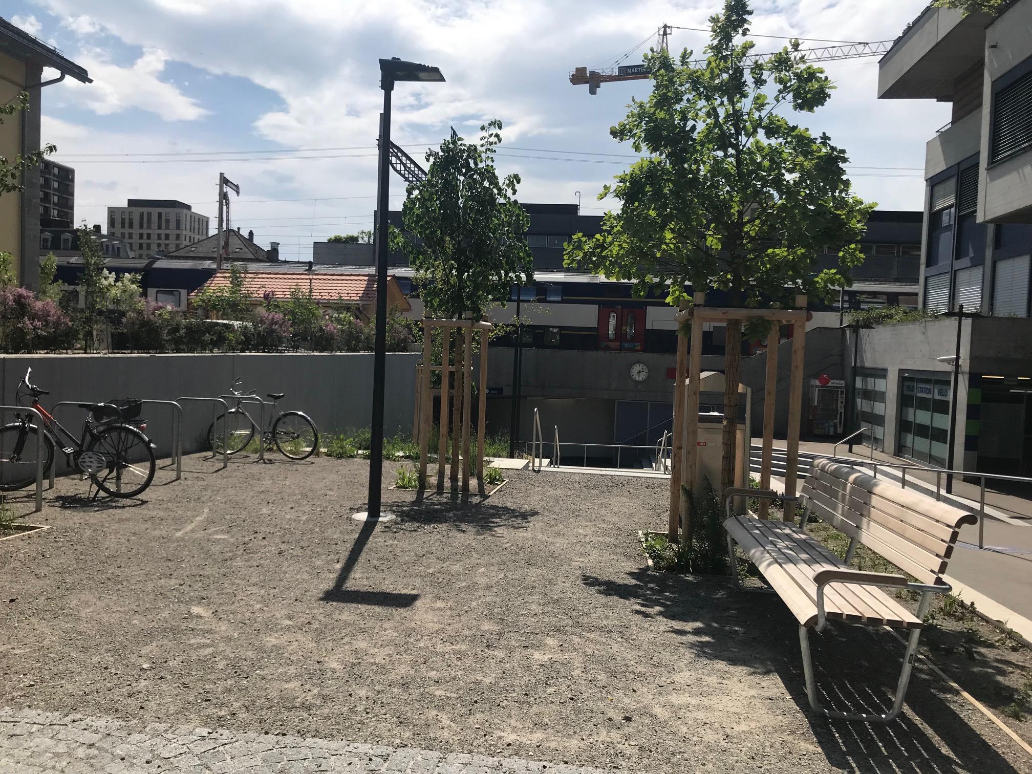 Der unversiegelte Platz direkt bei der Bahnhofsunterführung ist chaussiert. Es hat Sitzbänke, Veloständer und 2 neu gepflanzte Bäume. 
