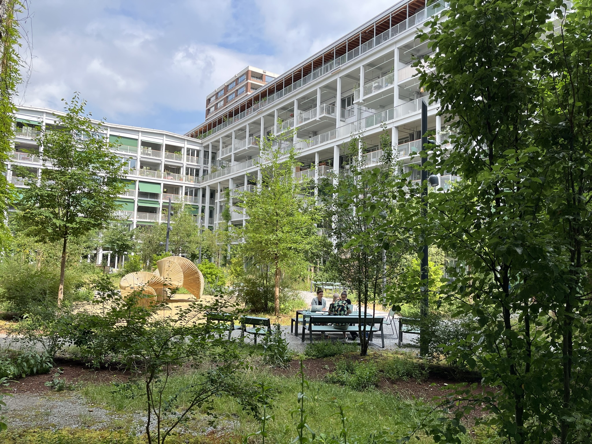 Der Innenhof der 6 bis 7 stöckigen Überbauung ist dicht begrünt. Der Boden besteht aus Kies und Gestrüpp.  Es hat viele mittelgrosse Bäume. An Sitzgelegenheiten mit Tischen sind Leute mit dem Laptop am Arbeiten. Ein Holzkonstrukt steht in der Mitte des Innenhofs und dient als Kunstwerk.