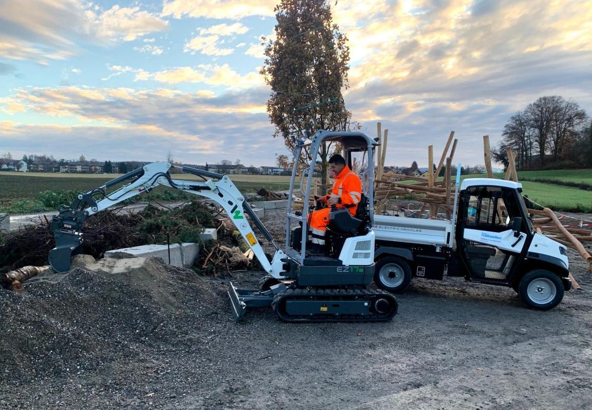 Ein vollelektrischer Kleinbagger trägt auf einer Baustelle einen Kieshaufen ab. Im Hintergrund ein kompaktes Elektro-Transportfahrzeug. 