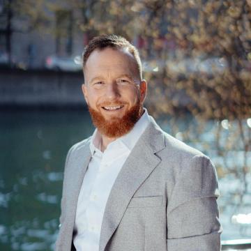 Steve Coucheman steht am Ufer der Limmat und trägt ein weisses Hemd und ein hellgraues Sakko. 