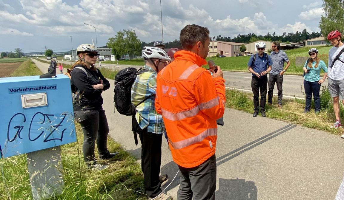 Eine Gruppe von Personen stehen auf einem Veloweg um eine hellblaue. kastenförmige Zählstelle für den Veloverkehr.