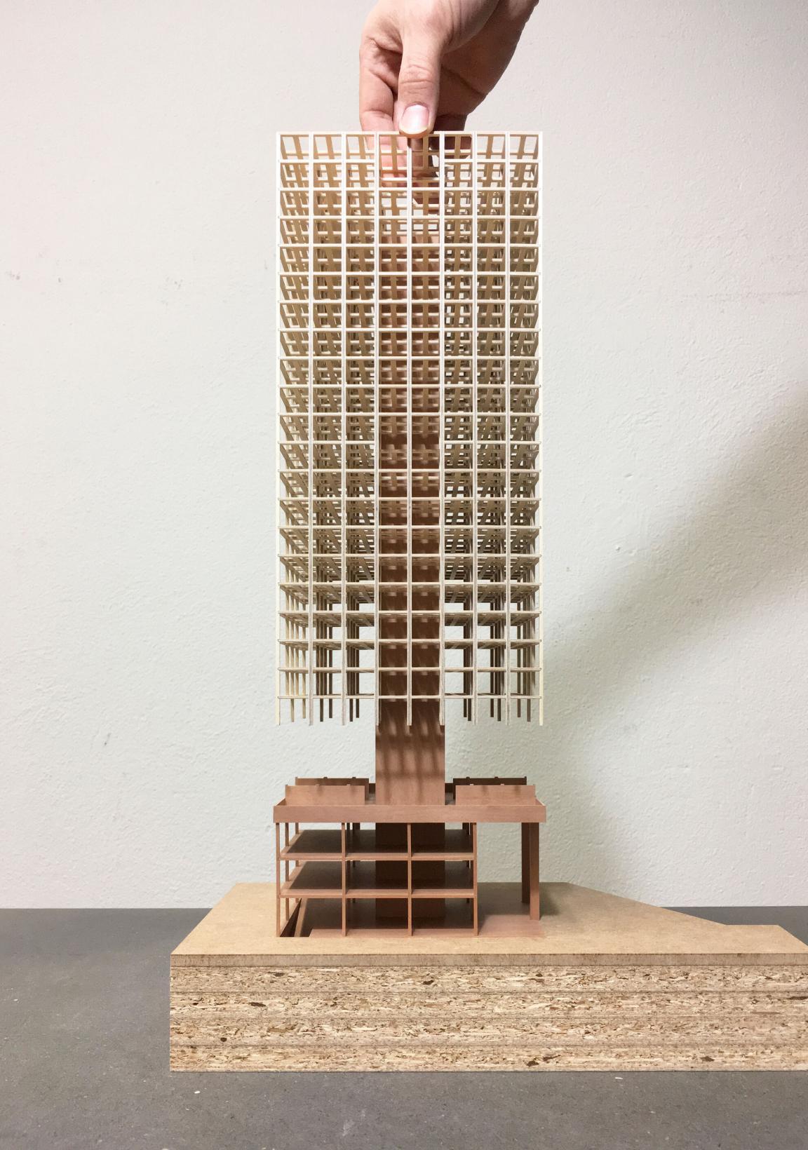 Modell des Holzhochhauses H1 mit Betonkern und Holzgitterfassade, von einer Hand gehalten, auf einer Holzplatte stehend.