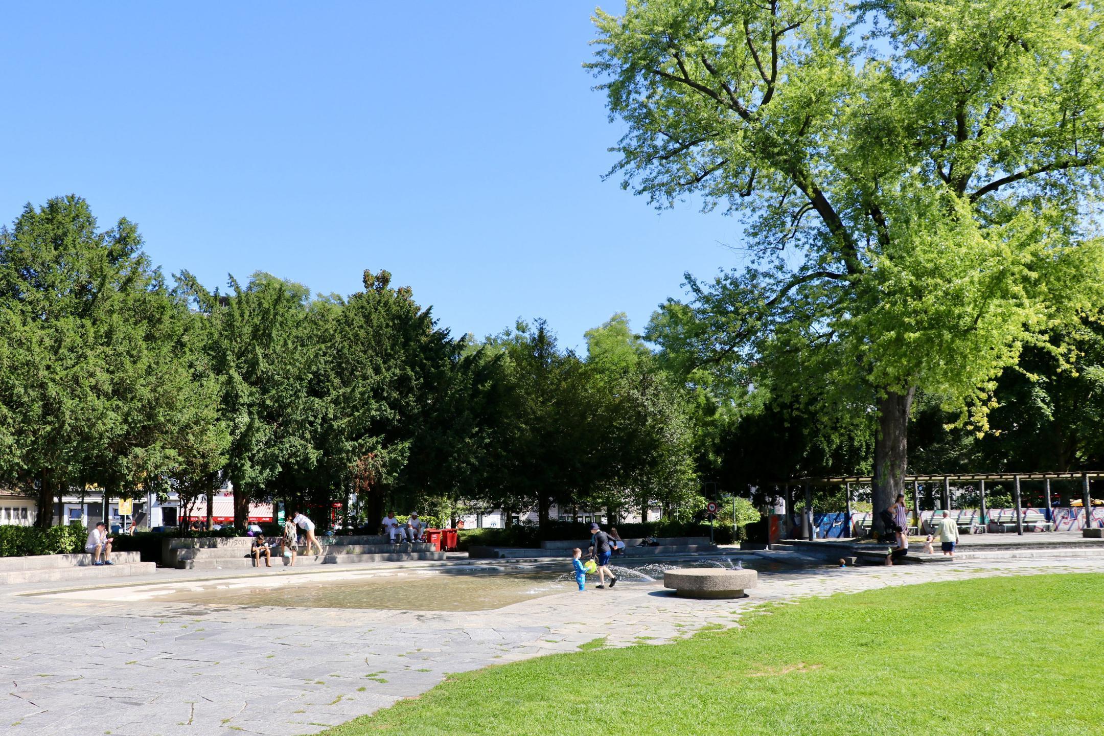 Wasserfläche neben Rasenfläche, welche teilweise durch Bäume beschattet wird, nutzen Kinder und Erwachsene als angenehme Abkühlung auf der Bäckeranlage in Zürich.