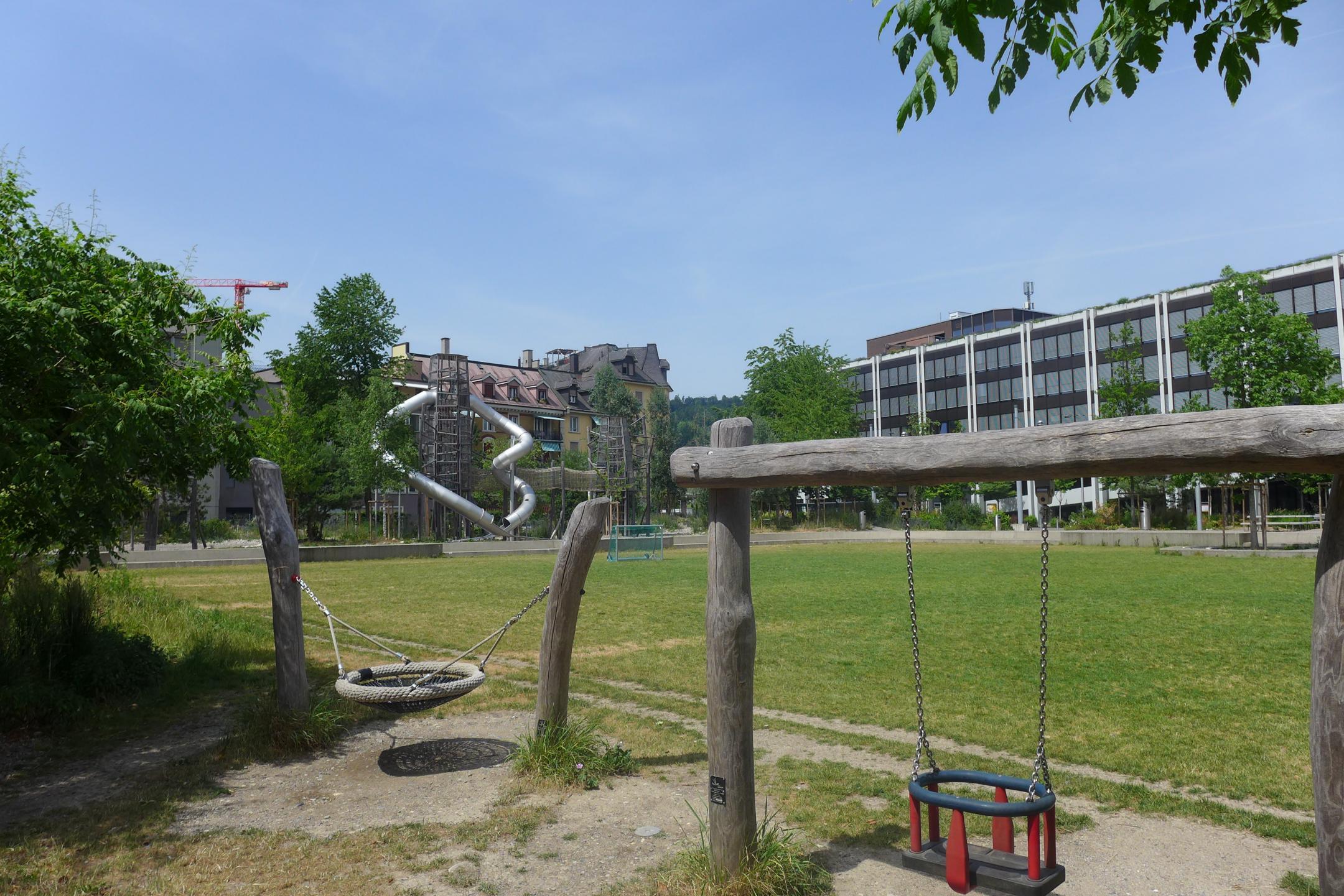 Spielplatz mit Rasenfläche des Quartieparks Schütze, welche von schattenspendenden Bäumen umgeben ist.