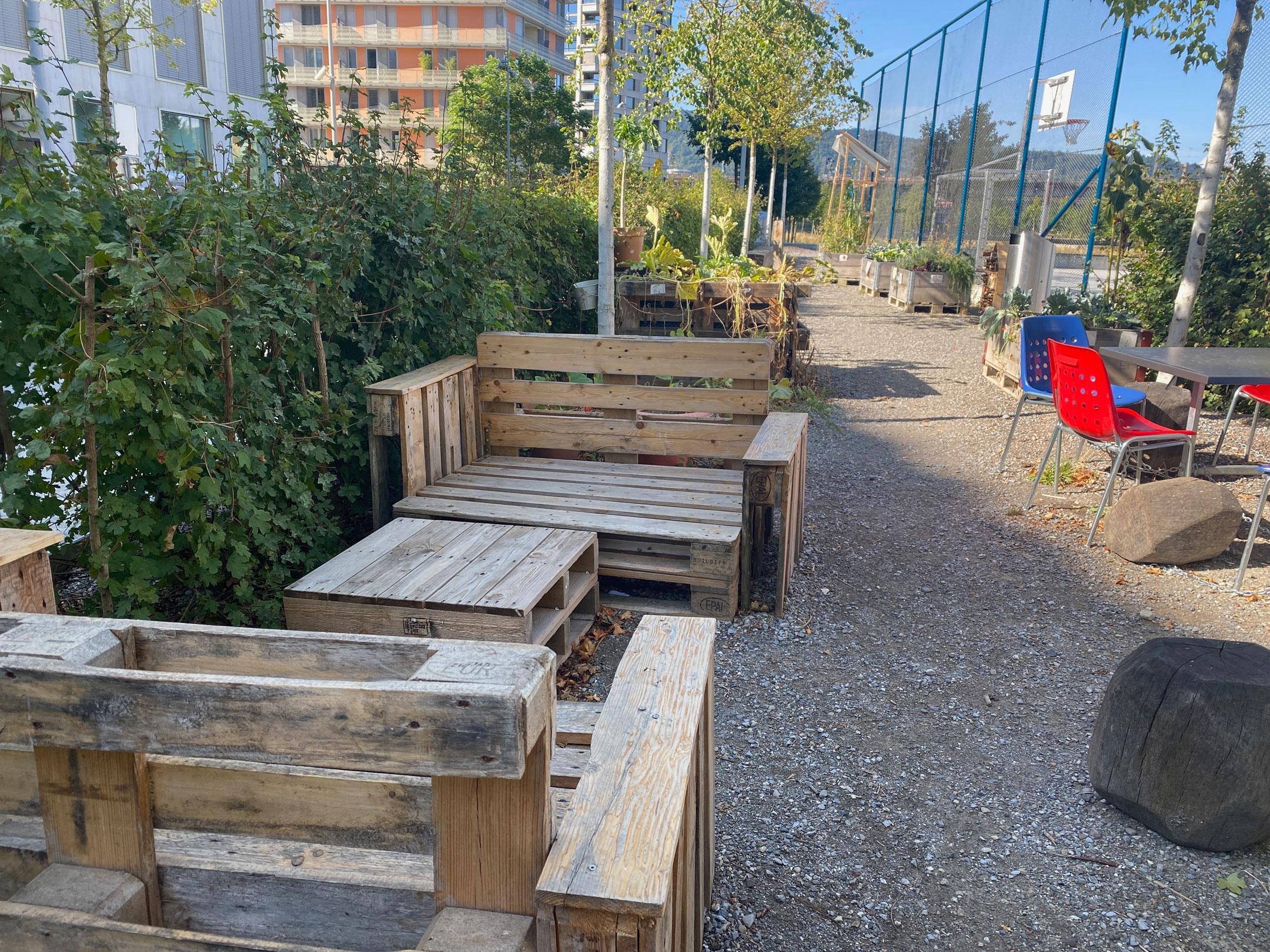 Das Foto zeigt einen Teil des Pausenplatze des Schulhauses. Auf dem Kiesboden stehen Tische und Stühle und eine aus Paletten selbst gemachten Sitzlounge. In Hochbeeten wächst Gemüse und es hat Bäume und Sträucher. 