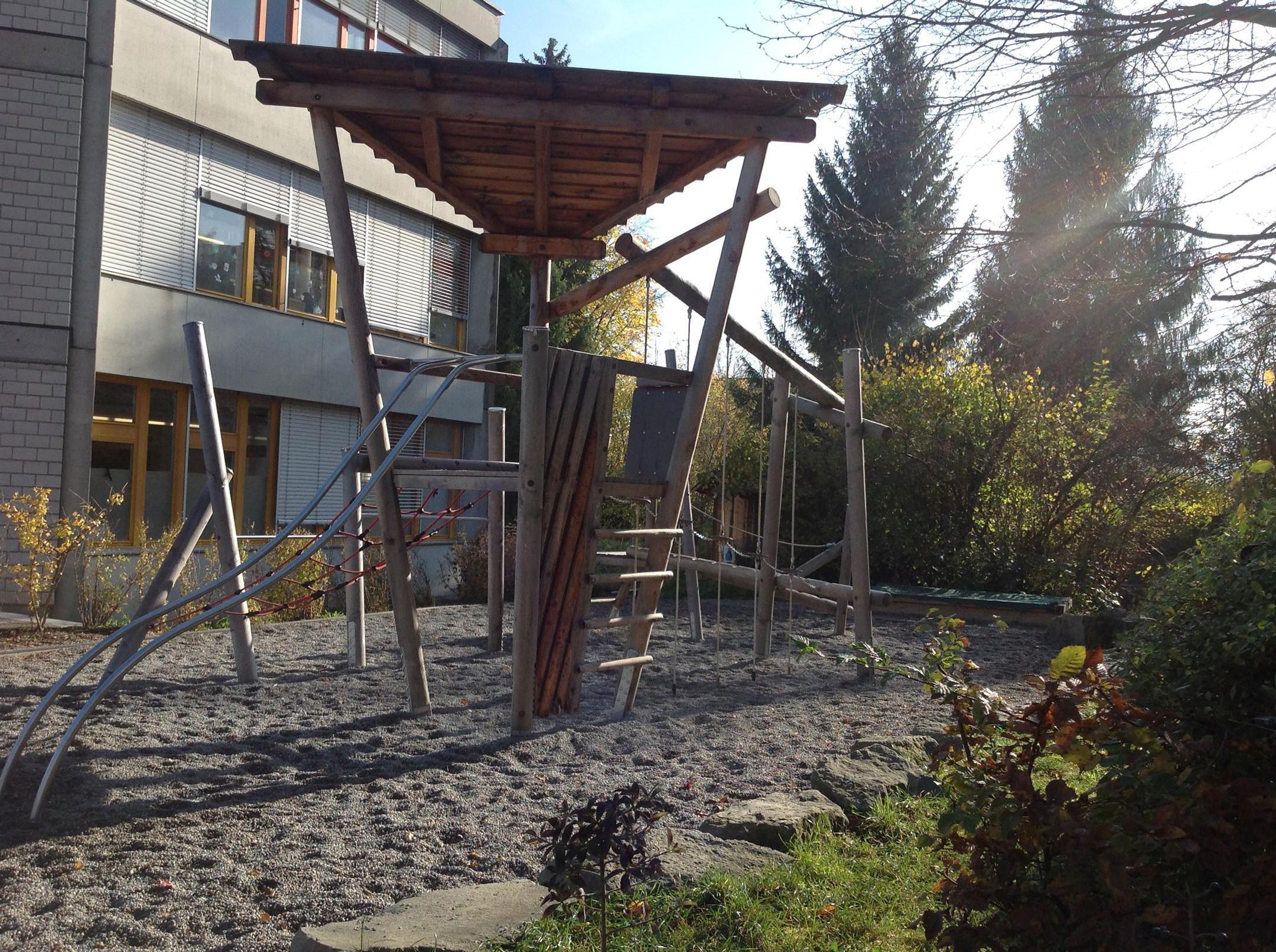 Foto eines Spielplatzes auf dem Pausenplatz des Schulhaus Zopf in Adliswil. Der Spielplatz zeigt ein Kiesplatz mit Spielturm aus Holz und viele gepflanzte Sträucher für die Biodiversität.