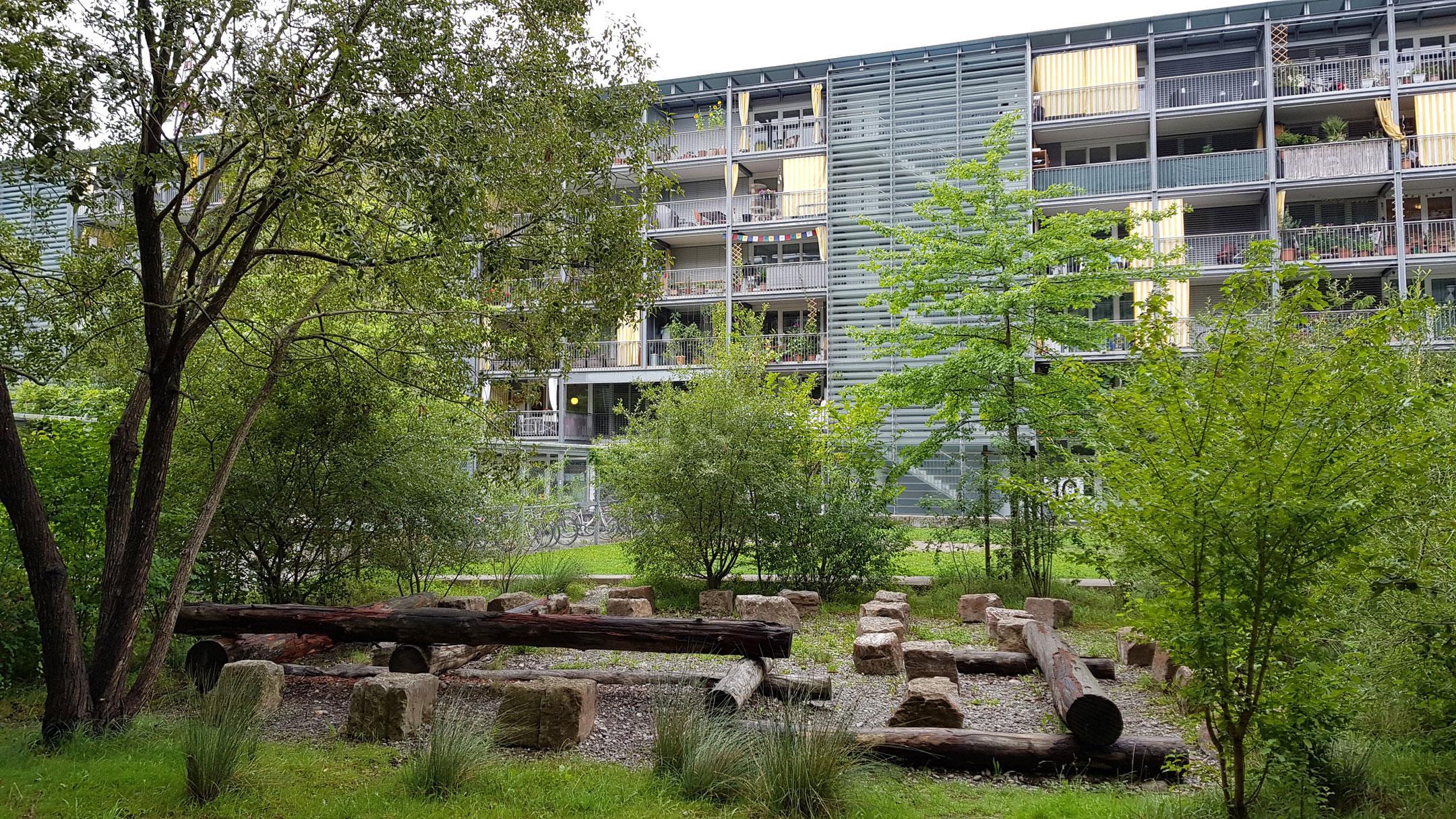 Das Foto zeigt einen Teil des Innenhofs der Regina Kägi Hof Überbauung in Oerlikon. Der Kiesplatz hat Baumstämme zum Sitzen oder drüber Balancieren und ist von Bäumen und Büschen umrandet.