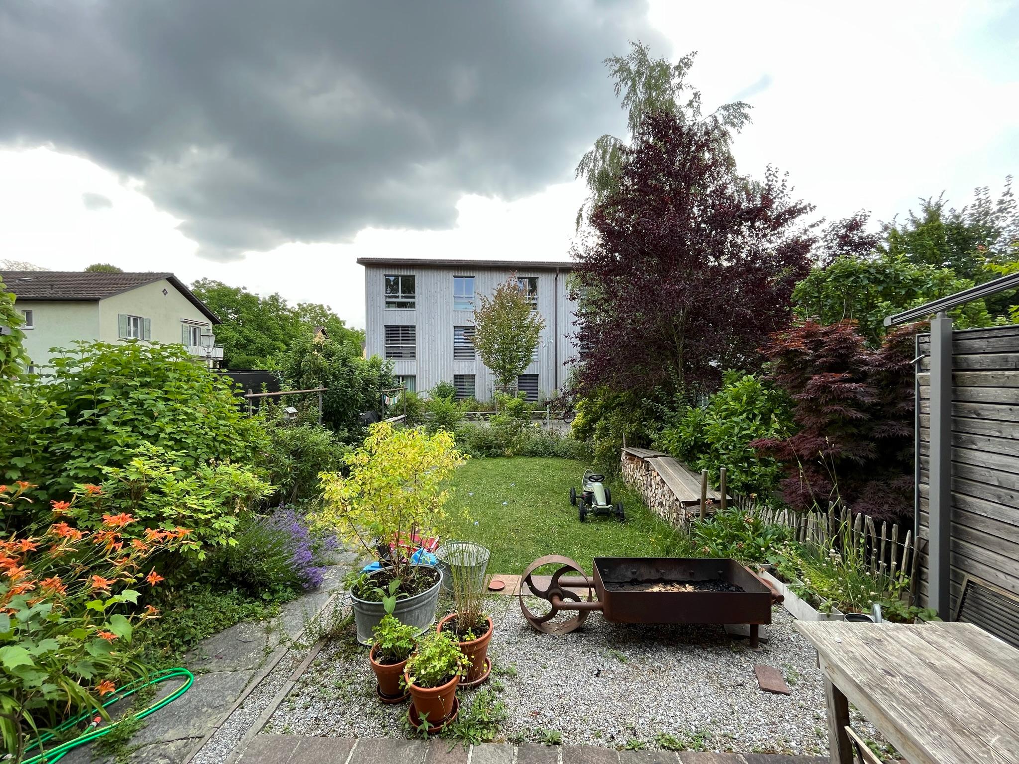 Das Foto zeigt den Garten eines Reiheneinfamilienhauses. Der Garten hat verschiedene Elemente wie Wiese, Kiesplatz und ein mit Steinplatten bedeckter Sitzplatz. Bäume, Sträucher, Blumen und eine Holzbeige umranden den Garten..