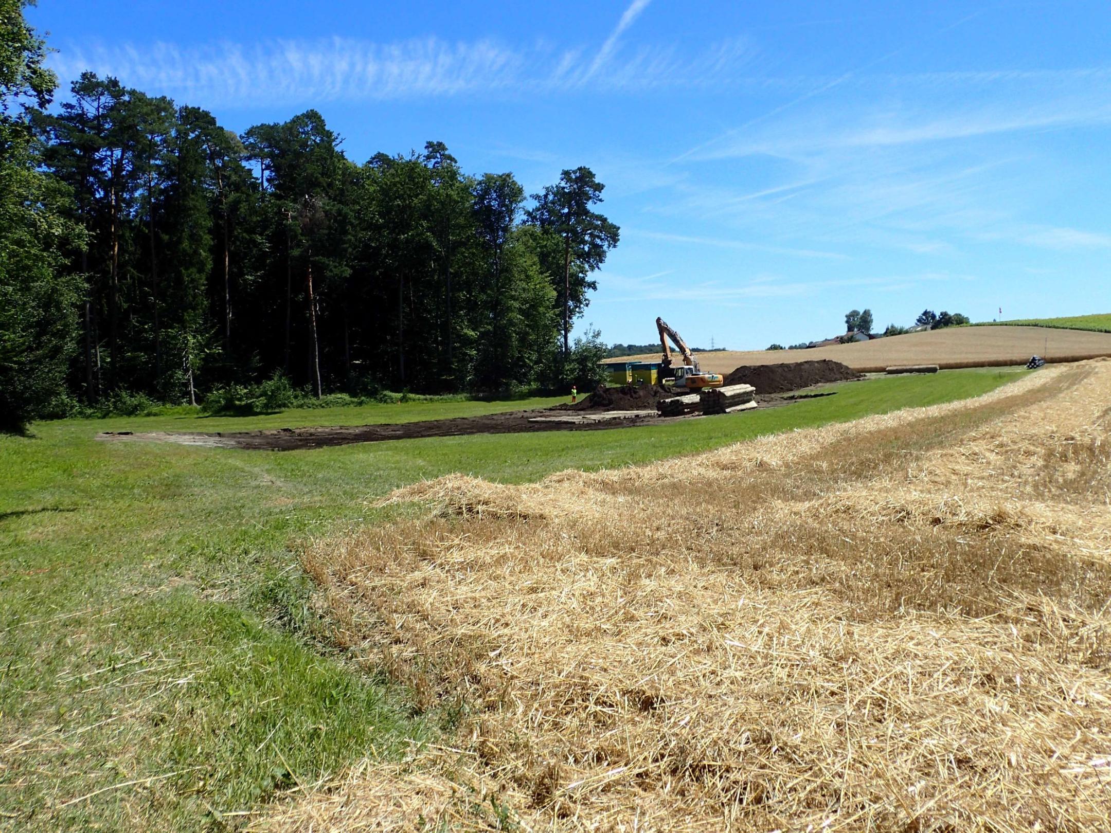 Im Vordergrund ist eine landwirtschaftlich genutzte Fläche. Im Hintergrund schaufelt ein Bagger Oberboden ab und nimmt Drainagen aus dem Boden, damit sich das Wasser dort zukünftig stauen kann.