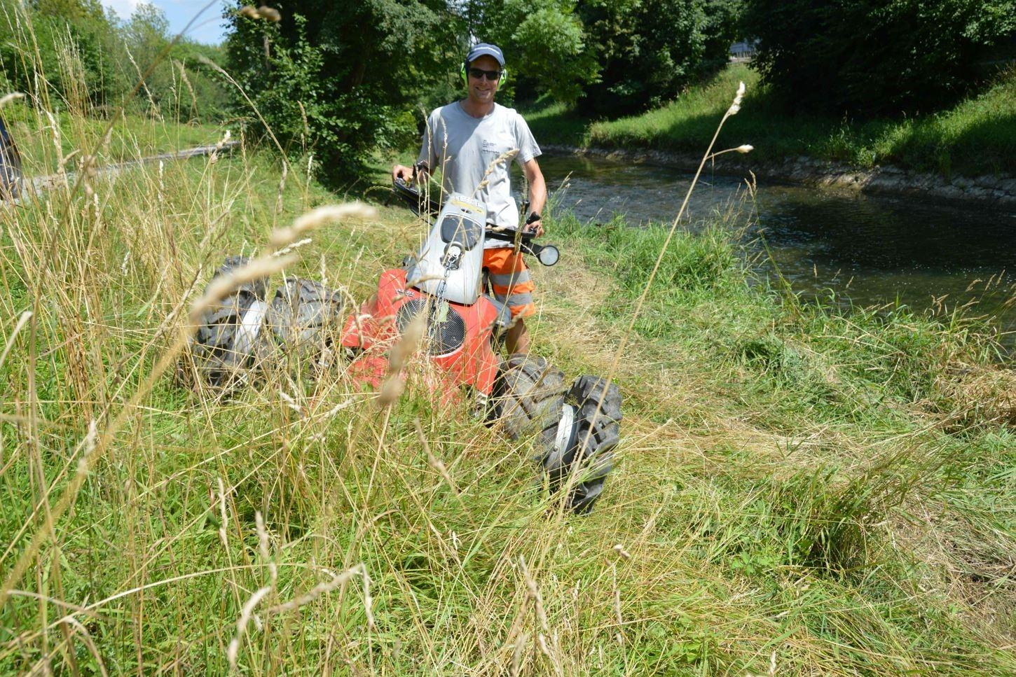 Auf einer leicht geneigte Uferböschung, die mit hohem Gras bewachsen ist, schiebt ein Mann einen Balkenmäher. Er trägt einen Hörschutz, Hut und Sonnenbrille. Hinter ihm sieht man das bereits gemähte Schnittgut am Boden liegen. 