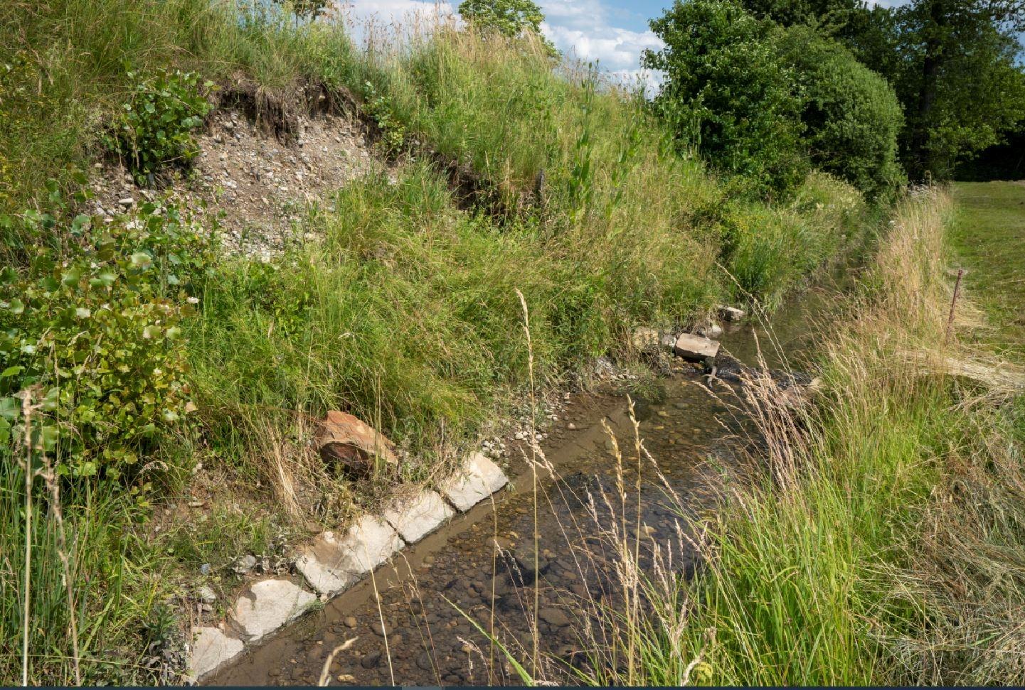 Blick auf einen begradigten, vollständig mit Mellonsteinen verbauten Bachlauf. An der bewachsenen, steileren Uferböschung links hat sich der Hartverbau in einem Abschnitt gelöst, wodurch ein Teil der Uferböschung abgerutscht ist. 