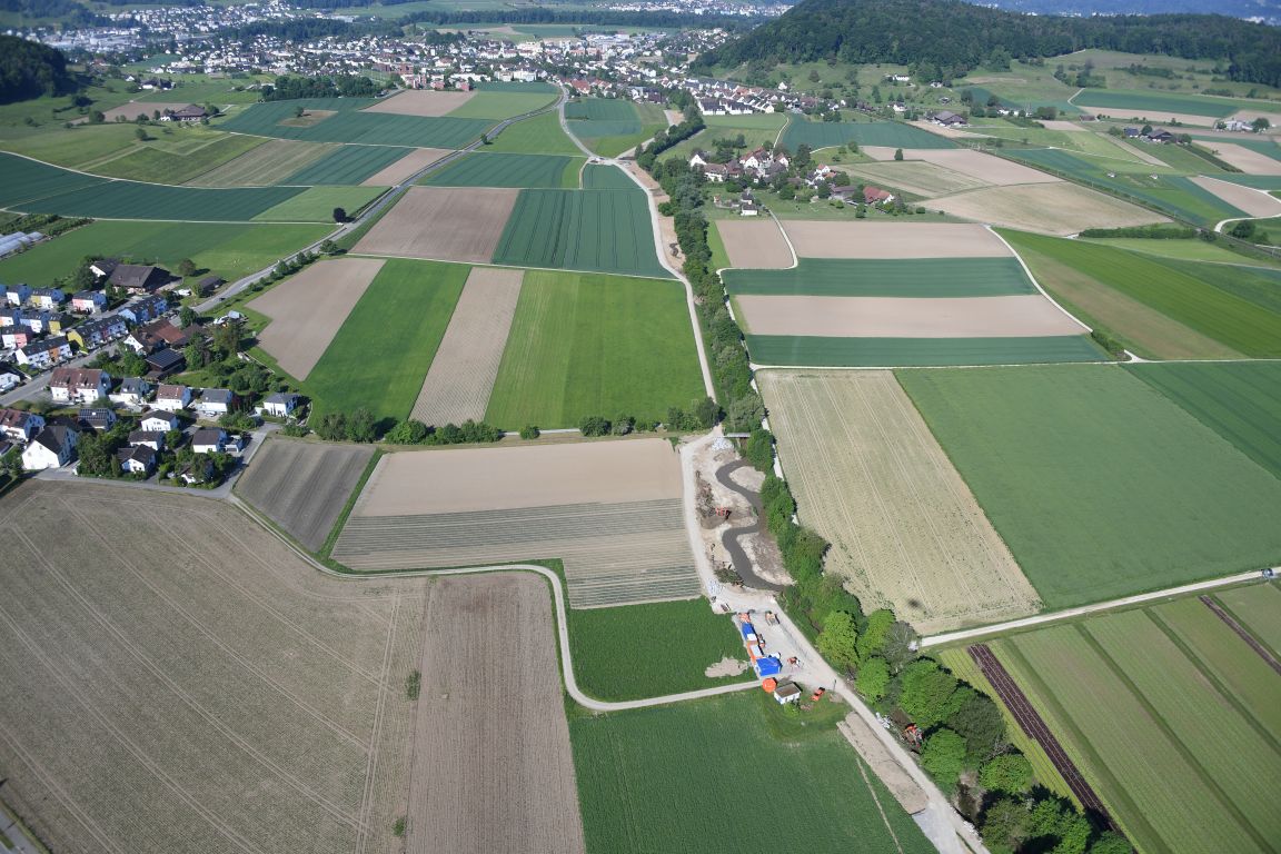 uftaufnahme einer ländlichen Landschaft mit Feldern und Gebäuden. Der Furtbach schlängelt sich durch die Landschaft, umgeben von Baumbestand und Ackerflächen. Ein Dorf mit weissen Häusern ist am linken Bildrand zu sehen.