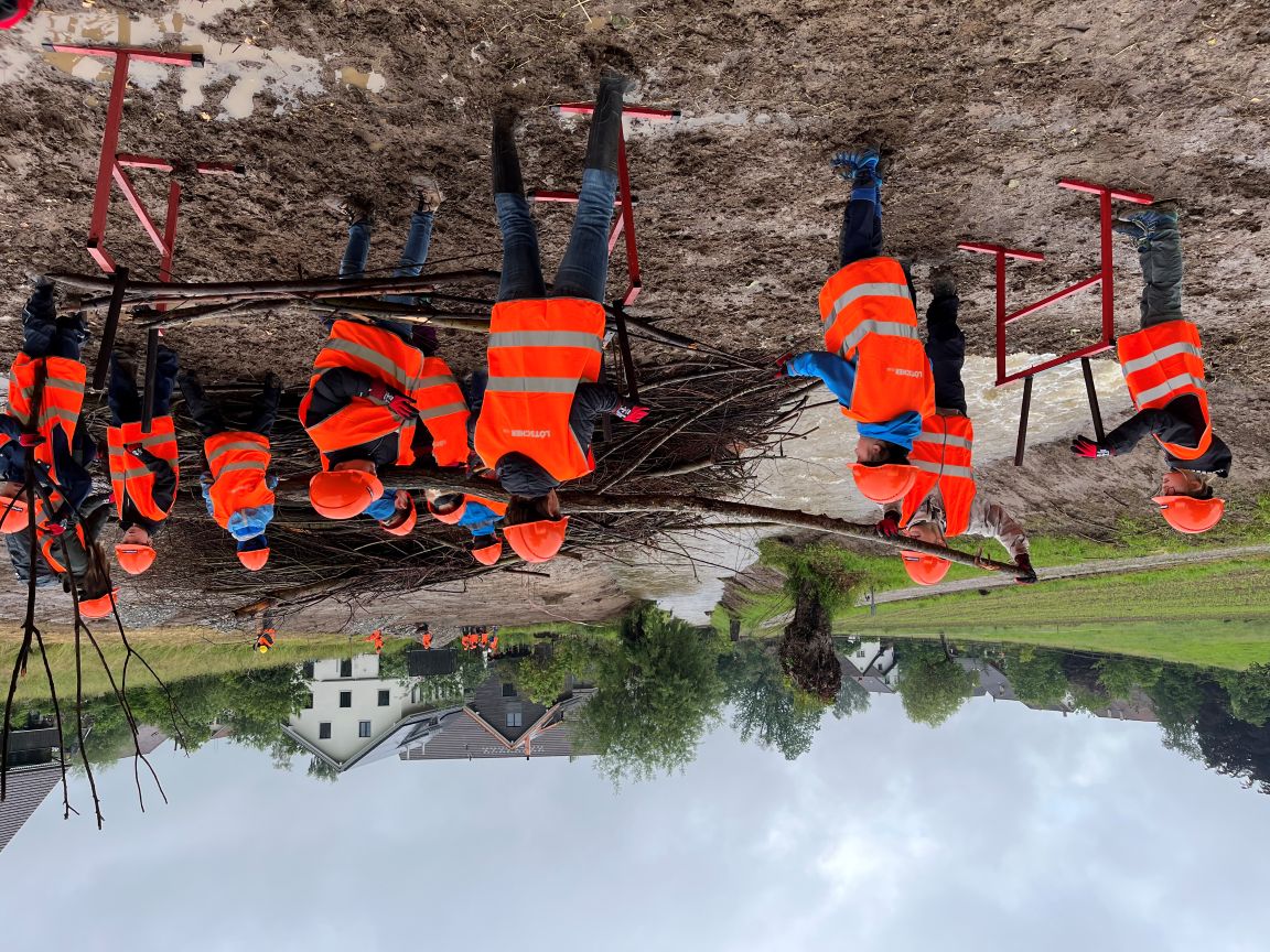 Schüler und Schülerinnen in Warnwesten und Schutzhelmen bauen mit Ästen und Schlamm einen Damm nahe eines Flusses, Häuser im Hintergrund.