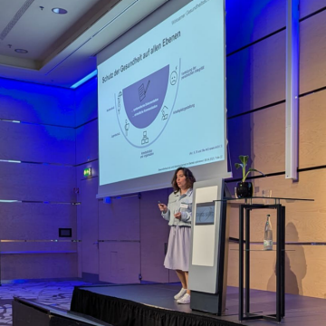 Milena Wütschert steht auf der Bühne und spricht das Publikum an. Hinter ihr zeigt eine Präsentationsfolie an der Wand die wichtigsten Ebenen des Gesundheitsschutzes an : Mutterschutz, Jugendschutz, Verletzung der persönlichen Integrität, Arbeitsstruktur und -organisation, Arbeitsplatzgestaltung