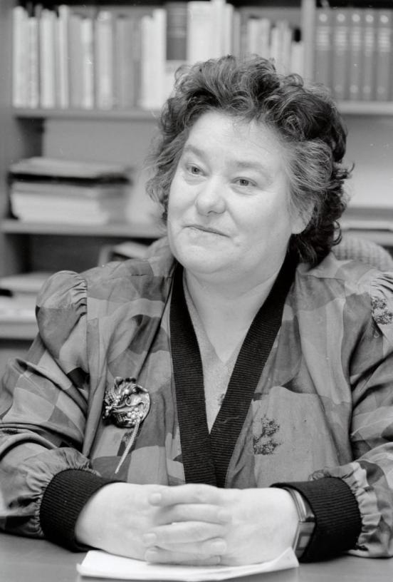 Eine schwarz-weiss Fotografie zeigt Hedi Lang mit verschärnkten Fingern an einem Tisch. Sie hat schwarz-graue Haare. Sie trägt eine grosse Brosche an der Kleidung. 
