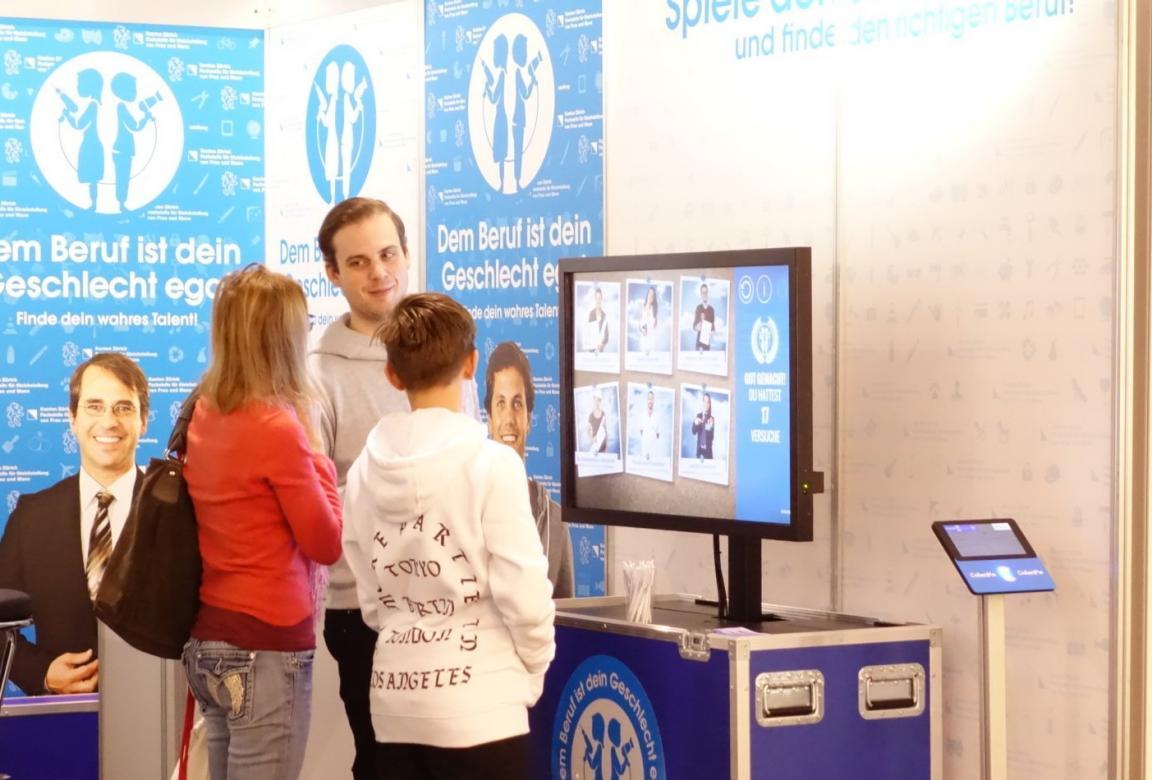 Ein Jugendlicher steht mit seiner Mutter an meinem Stand der Berufsmesse. Sie schauen auf einen Bildschirm. Am Stand steht ein Mann, der mit ihnen spricht. Der Jugendliche hat kurze Haare und einen weissen Pullover. Die Mutter hat blonde Haare und einen roten Pullover. Der Mann am Stand trägt kurze Haare und einen grauen Pullover.