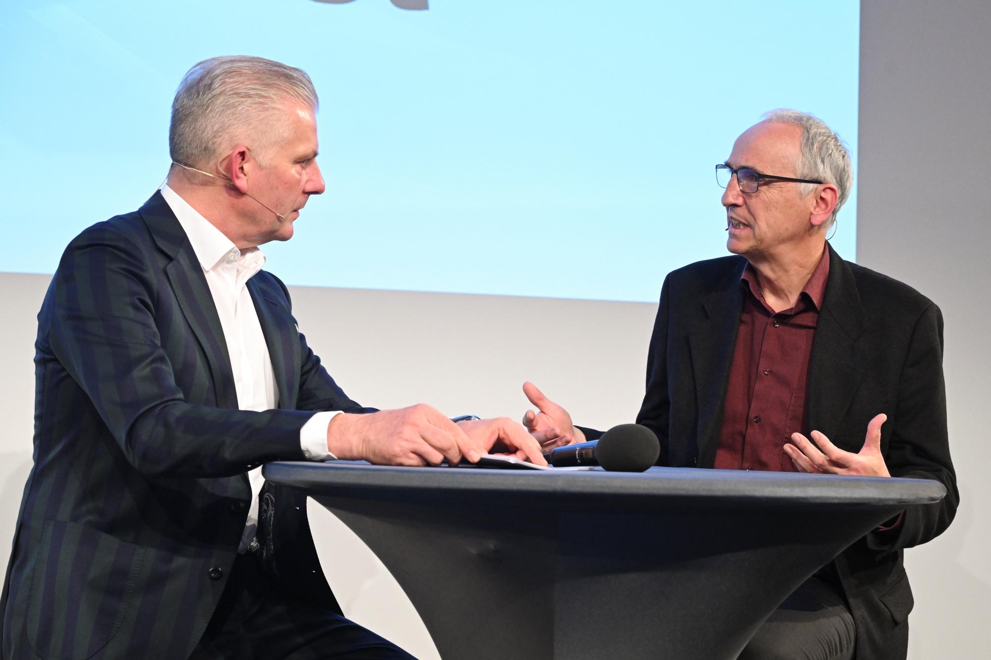 Markus Herren und Rainer Maria Salzeber im Gespräch an einem kleinen Tisch auf der Bühne.