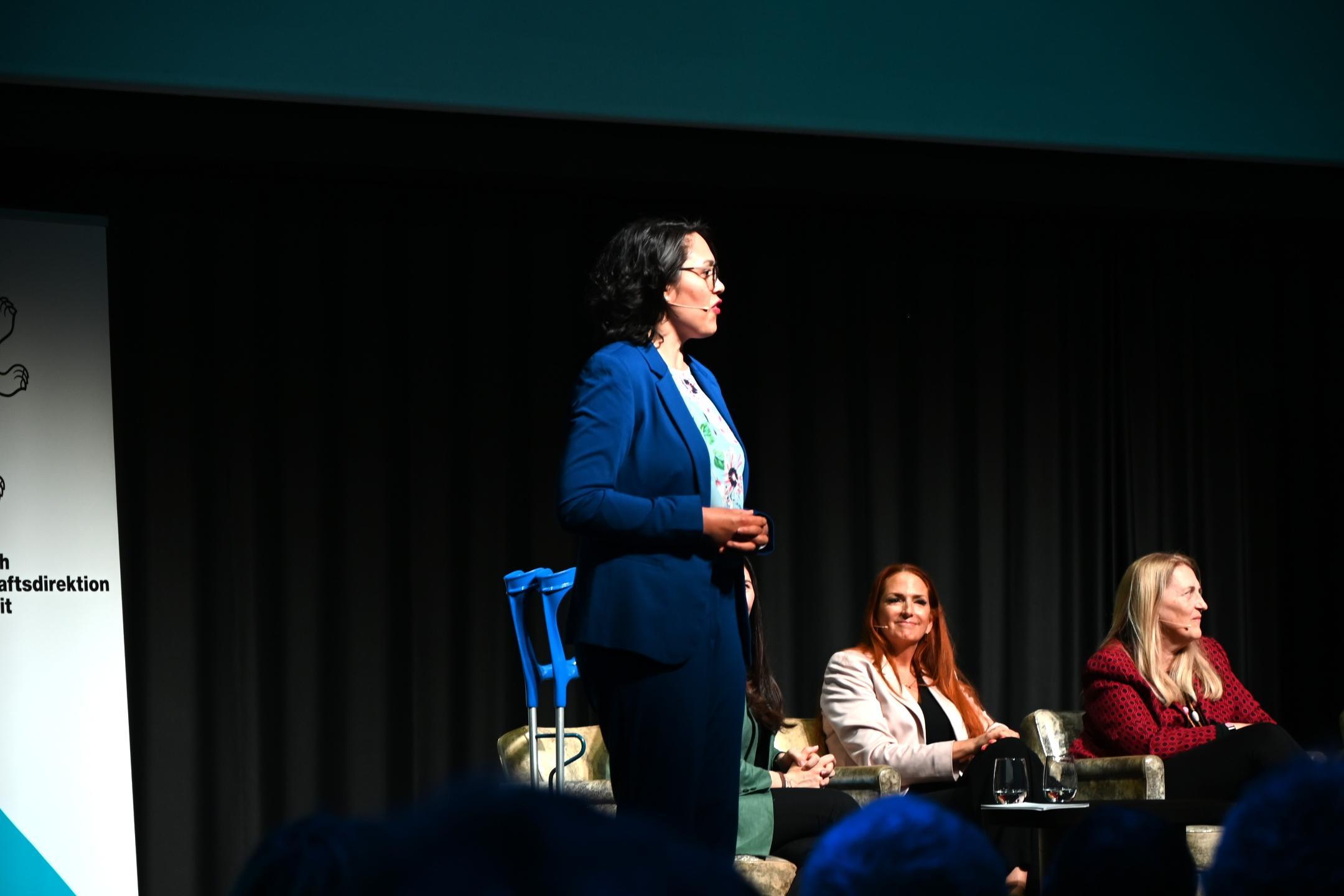 Joy Hofstetter, Fachverantwortliche des Mentorin-Programms, steht auf der Bühne. Im Hintergrund sind die Referierenden zu sehen