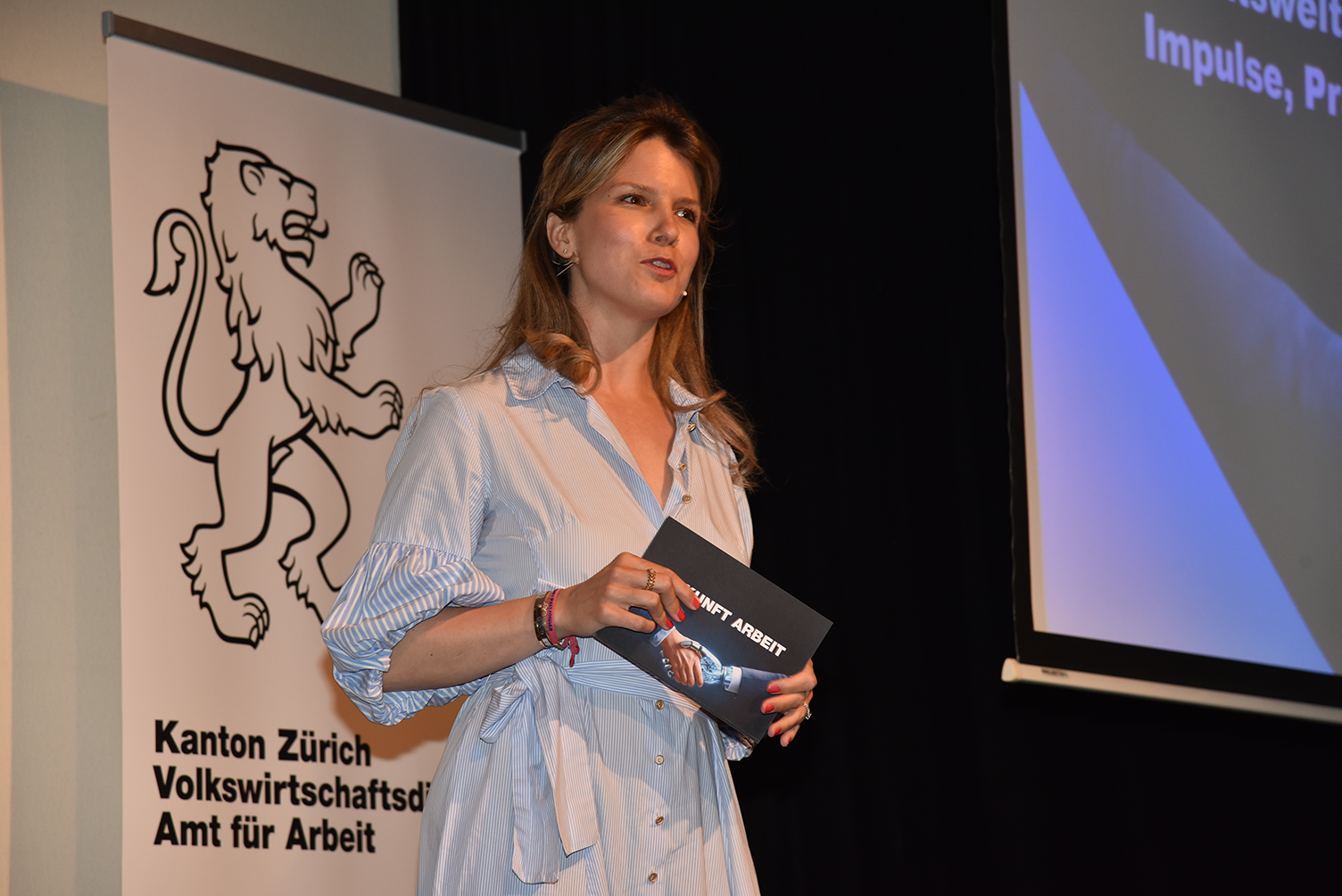 Moderatorin Valérie Henzen begrüsst das Publikum. Hinter ihr sieht man das Logo des Amts für Arbeit.