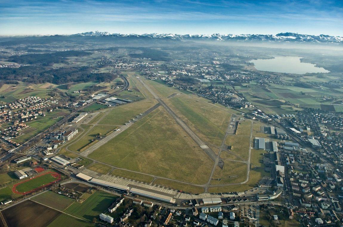 Eine 2,8 km lange Piste des Militärflugplatzes von oben, umgeben von Wiesen und Gebäuden. Am oberen Bildrand sind Schneebedeckte Berge zu sehen und darunter ein See.