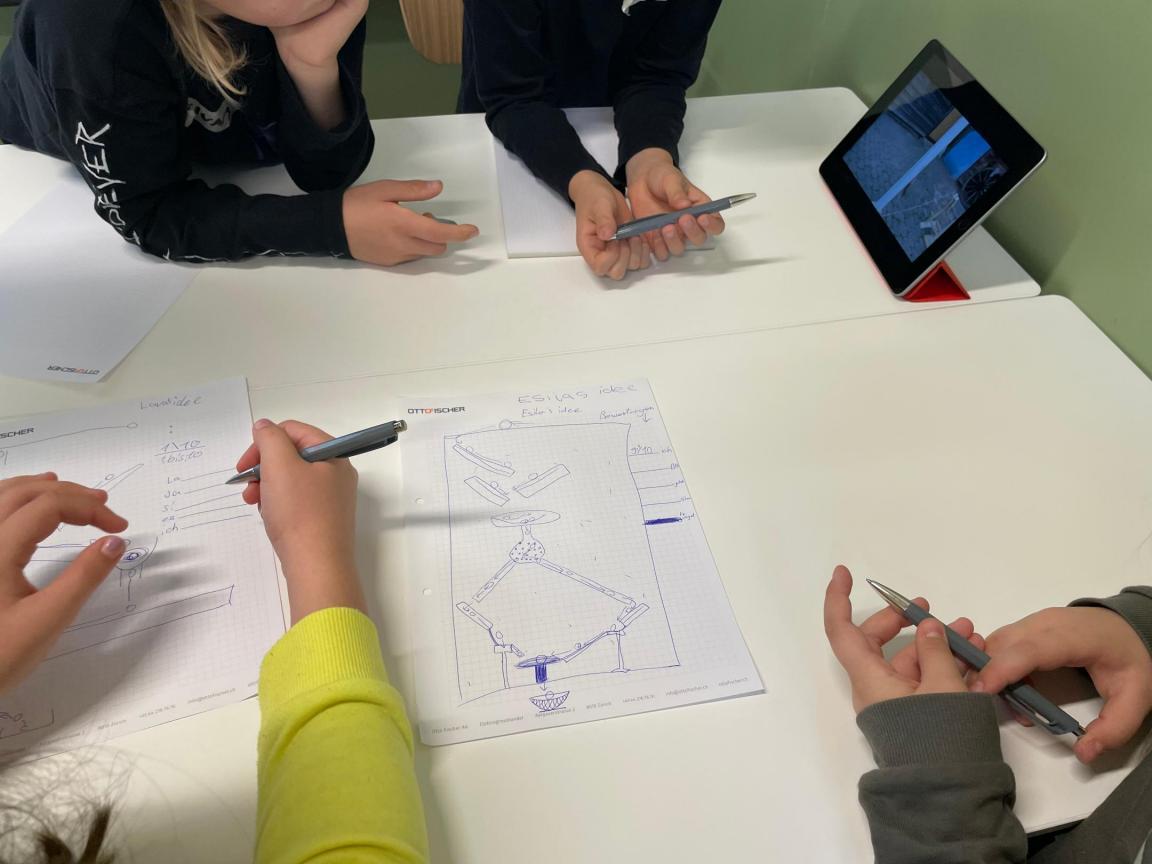 Ansicht von schräg oben auf einen Tisch an dem Kinder sitzen und an einer Schulaufgabe arbeiten
