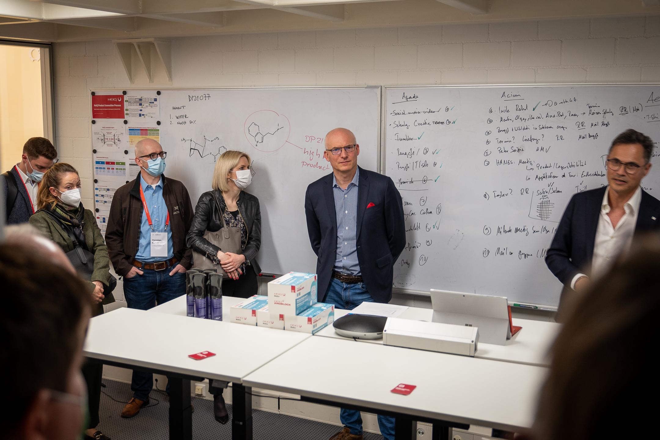 Personen stehen um Tische herum und hören jemanden zu