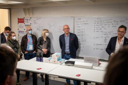 Personen stehen um Tische herum und hören jemanden zu